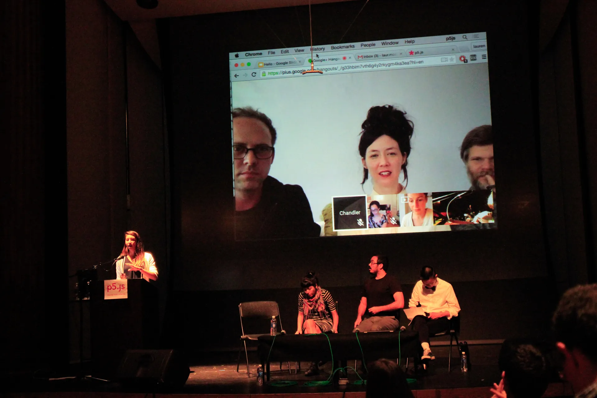 Woman speaks at a podium in an auditorium while three participants sit on the stage and another three are skyping in on the stage screen