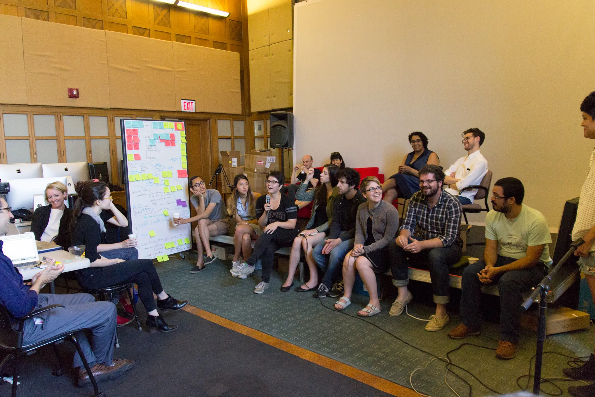 Participants sit in a circle around a white board with sticky notes on it while a female student speaks into a microphone