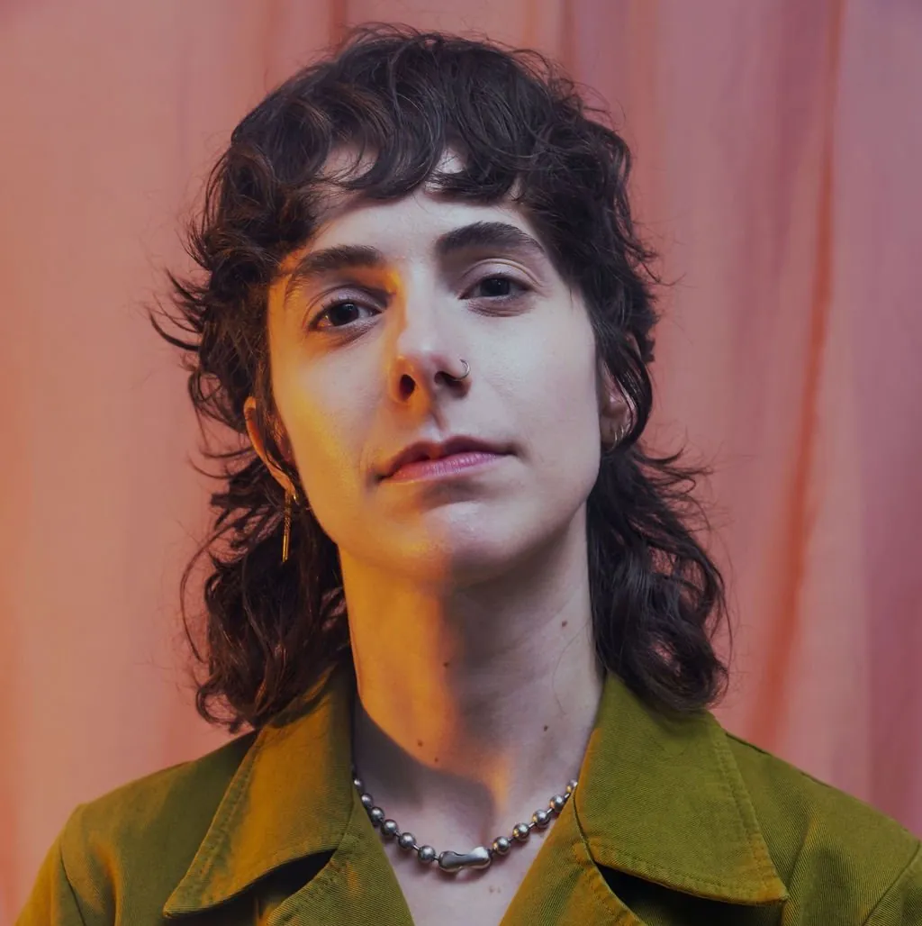 Headshot of Cassie Tarakajian, a light-skinned Armenian-American non-binary person with a neutral face, with a brown mullet, wearing a green shirt against a mauve background.