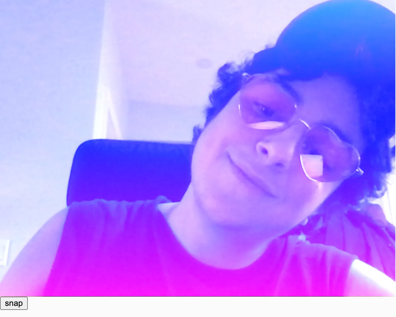 A white person with short dark curly hair, a dark sleeveless t-shirt, and pink heart shaped glasses tilts his head and looks down at the camera, smiling softly. A blue to pink vertical linear gradient is overlaid on top of the photo.  Because of the LIGHTEST blendMode we're using, the colorful gradient is especially visible on his dark hair and dark shirt.