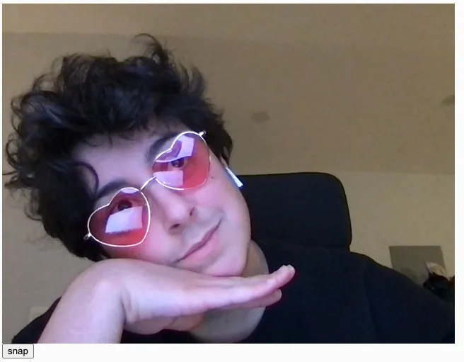 A white person with short dark curly hair wearing pink heart shaped glasses posing with one hand under his chin, softly smiles, and looks to the right.