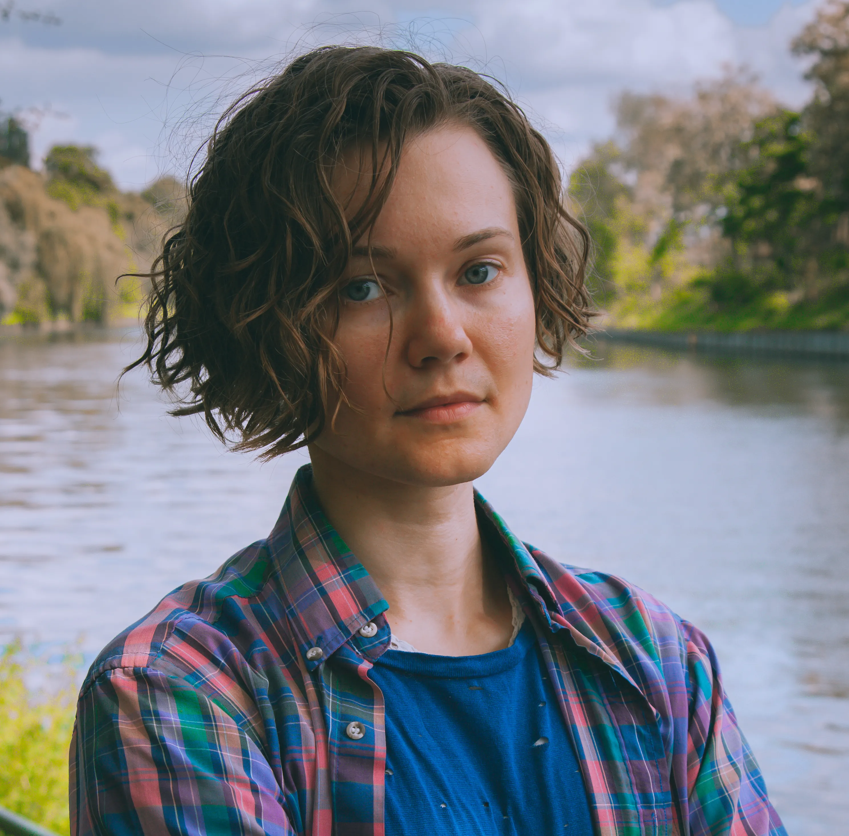 A photograph of a white non-binary person with short blonde hair. They stand against a background of a canal and greenery.