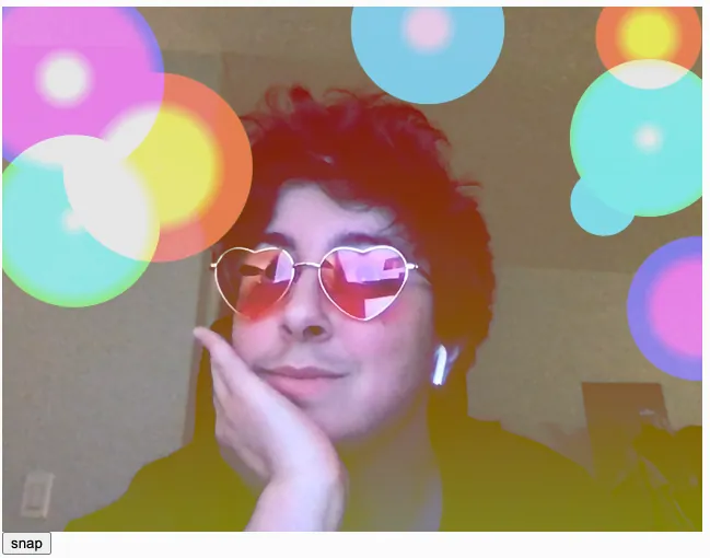 A webcam snapshot of a white person with short dark curly hair wearing pink heart shaped glasses posing for a picture. There is a red to yellow linear gradient filter on top of the photo. Colorful circles are scattered on top of the image with a color gradient overlay. The circles have randomized radial color gradients inside them to mimic a camera's lens flare. Some color gradients are pink to purple, cyan to green, or yellow to orange.