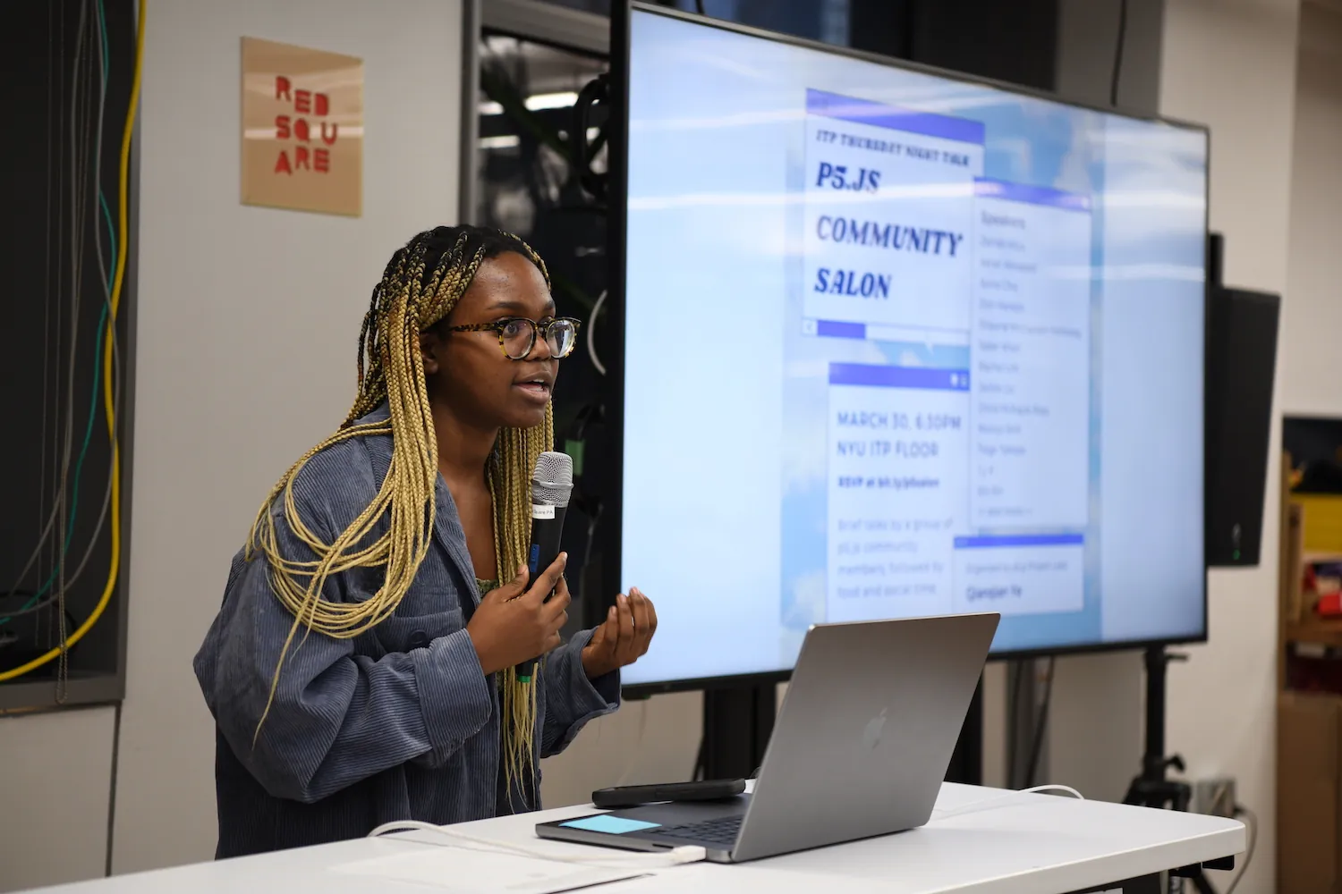 Olivia Ross holding a mic next to a big t.v. screen that has the p5 salon poster on it.