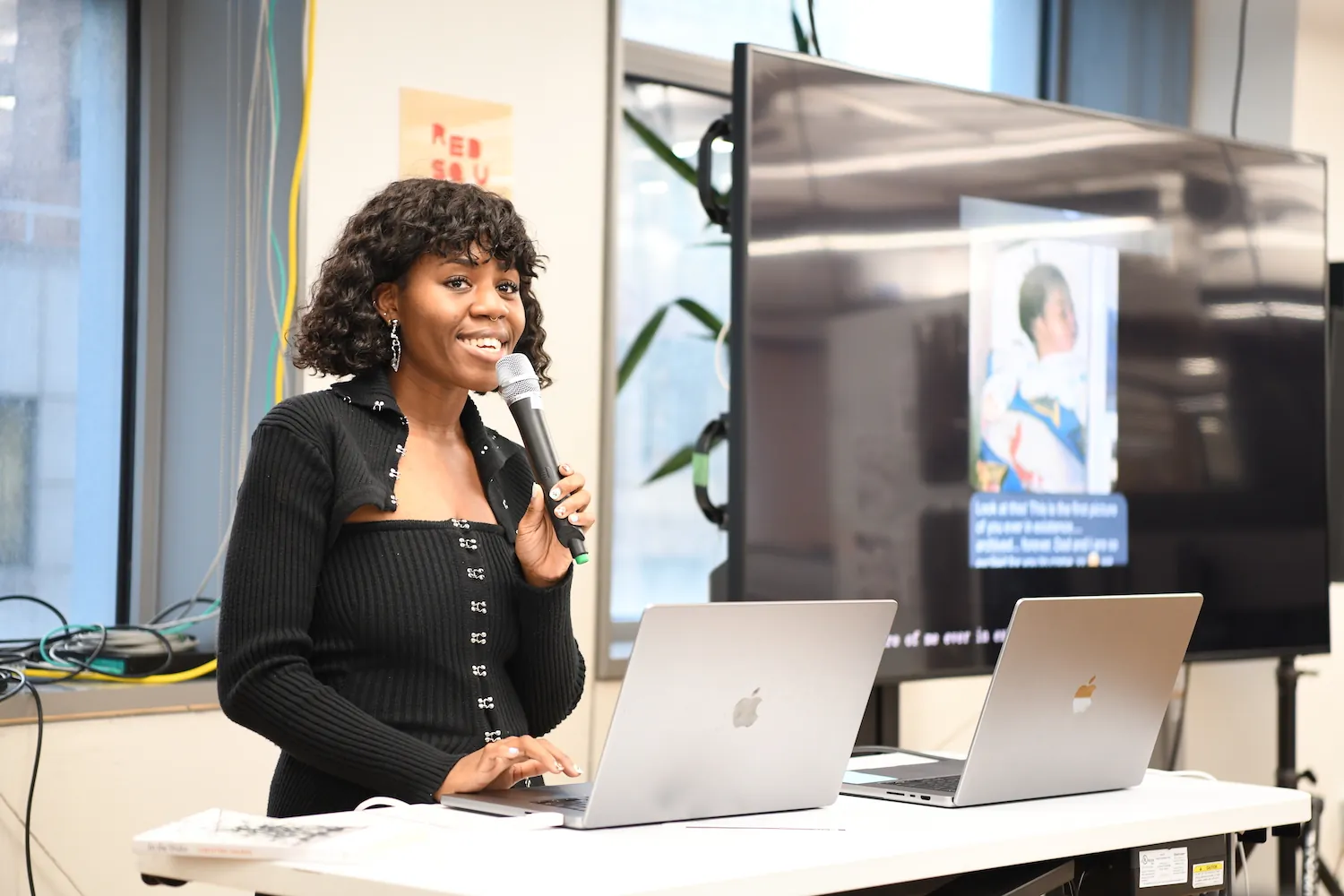 Zainab Aliyu holding a mic next to a big t.v. screen that has an image of her work. She is holding a book and reading and excerpt from it.