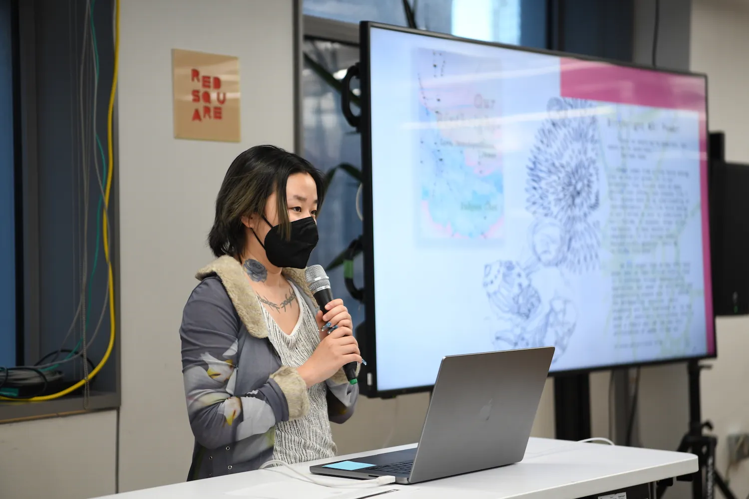 Suhyun Choi holding a mic next to a big t.v. screen that has an image of their work.